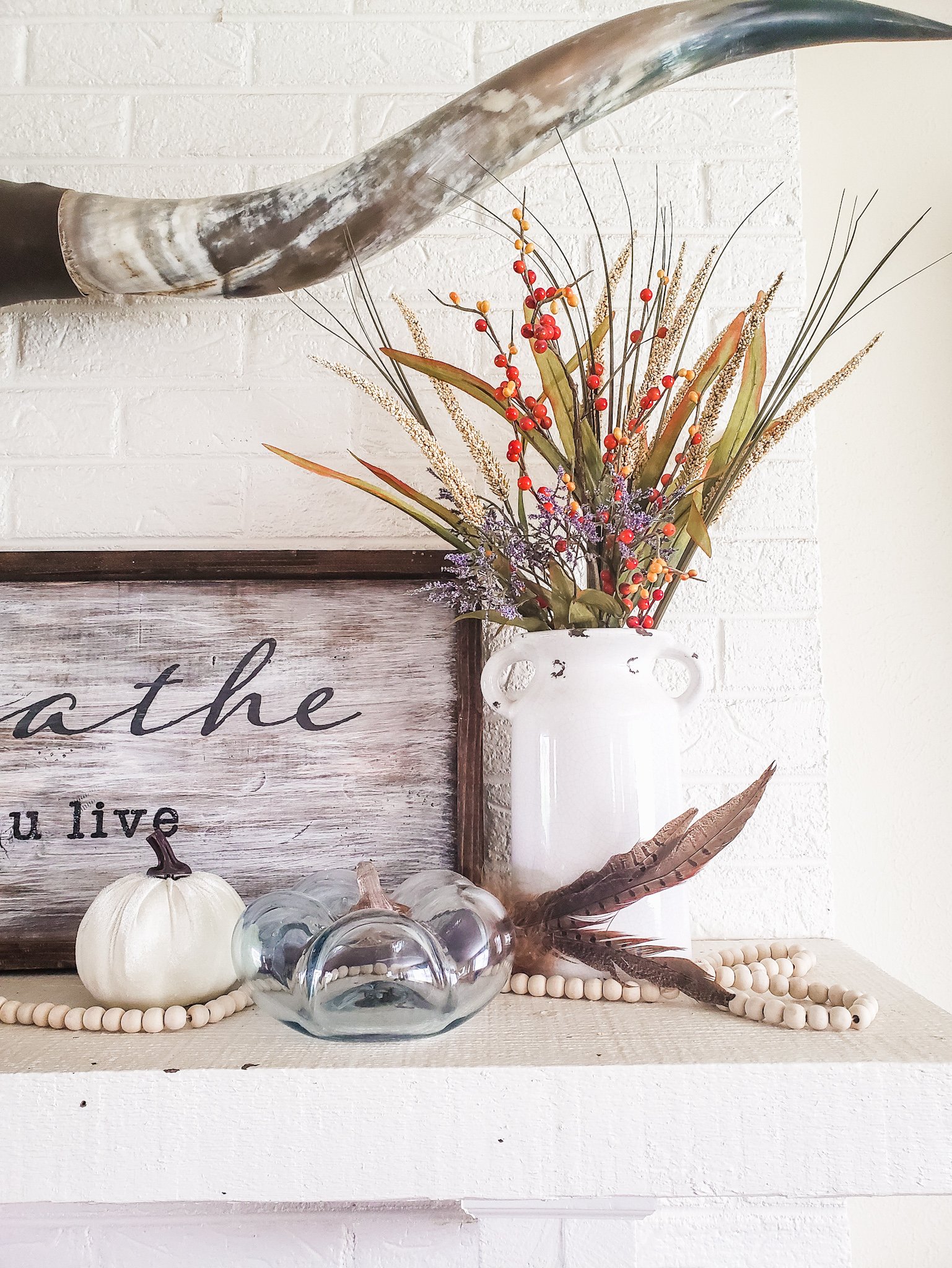 closeup of a fall mantle showing mounted longhorns, wooden sign, velvet pumpkin, glass pumpkin, pheasant feather, wood beads and a milk jug with autumn foliage.