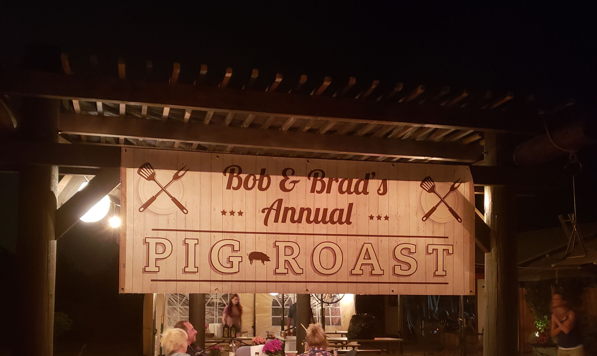 Pig Roast sign hanging from a pergola. People sitting at a table under the pergola enjoying their food.