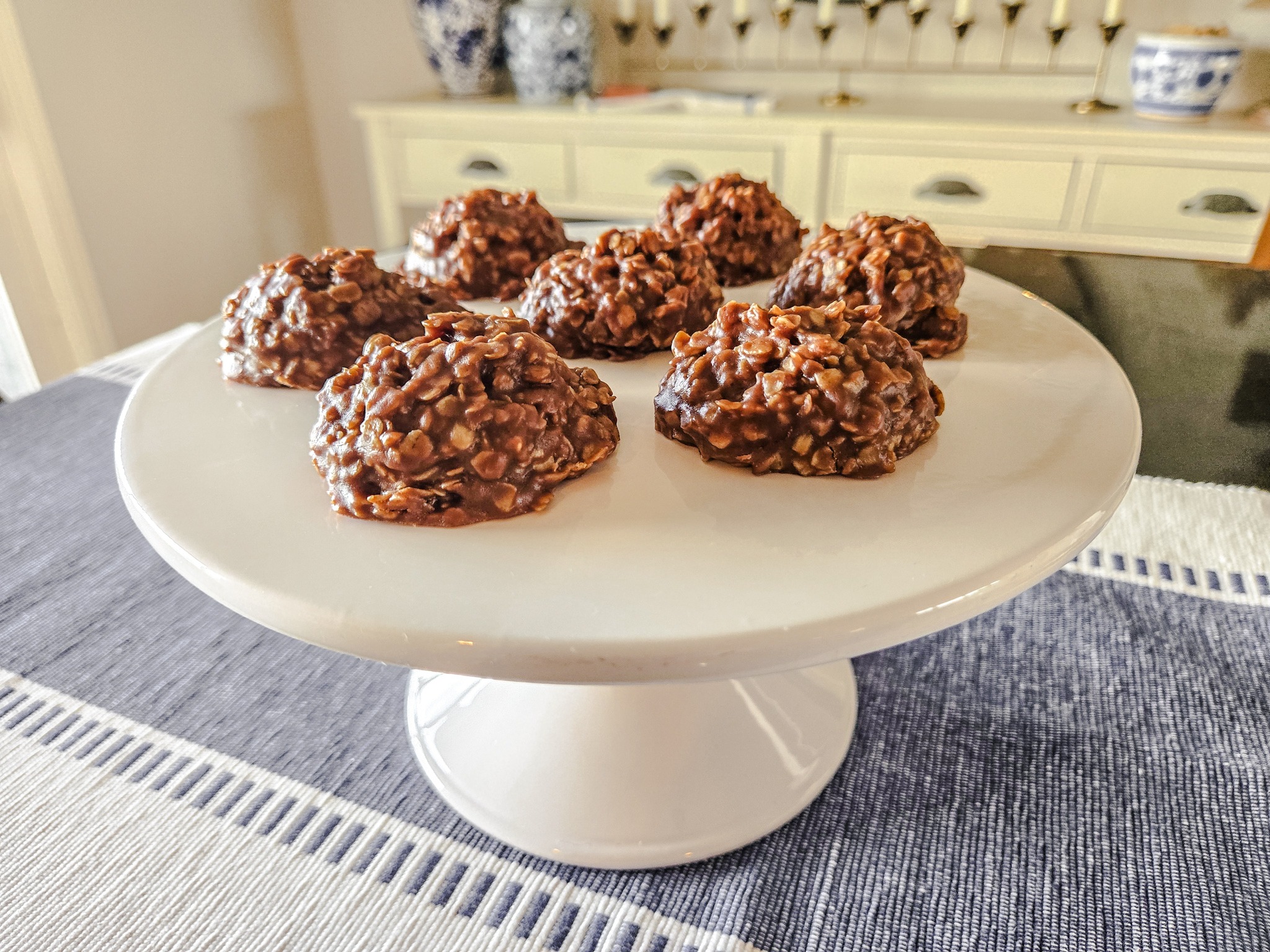 cookies on a white pedestal on a kitchen table with a blue and white table runner.